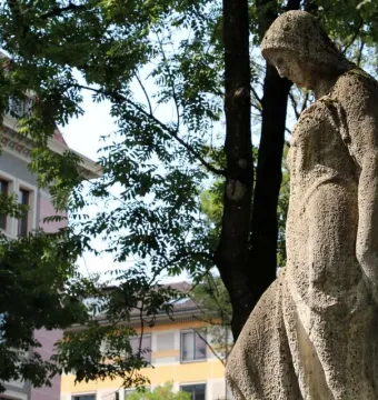 Weathered stone figure from the Nornenbrunnen stands beside trees and colorful buildings in the background.