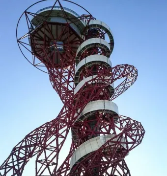 Orbit Tower’s red metal framework with a white spiral walkway rising to the top against a blue sky.