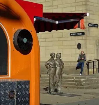 The Women of Steel sculpture stands outdoors on a paved area near a stone building, with street signs and a person in the background.