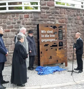 Rusted Dejudaization Institute memorial with German text and abstract cut-outs, surrounded by several people during an outdoor unveiling.