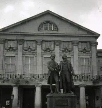 Goethe and Schiller Monument on a pedestal in front of a classical building, with people gathered in the square.