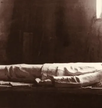 Käthe Kollwitz\'s lying dead soldier sculpture shown full length in military uniform against a dark wall.