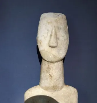 Kykladic Idol marble figurine shown from the upper torso up, with a long neck and stylized head against a dark background.