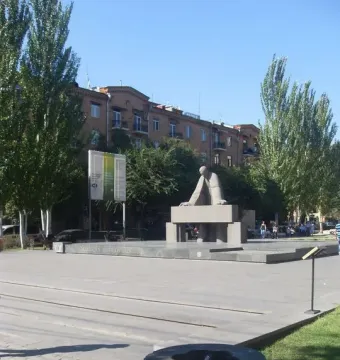 Monument to Alexander Tamanyan on a paved plaza with trees, buildings, and a stone fountain nearby.