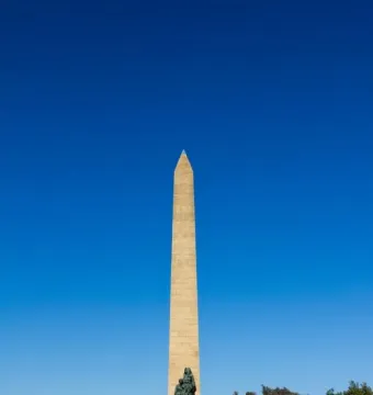 National Women\'s Monument obelisk with a bronze woman and child sculpture at the base, framed by trees and shrubs.