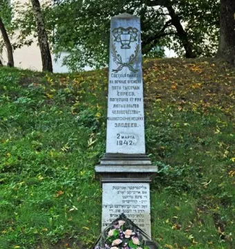 The Pit monument, a tall grey stone memorial with inscriptions and a carved emblem at the top, stands on a grassy hill with wreaths and flower baskets at its base.