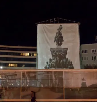 Tsar Liberator Monument covered by a large white banner on scaffolding at night, with nearby buildings and street activity.
