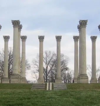 Capitol Columns standing in a wide arc on a grassy field with steps in the foreground and cloudy skies above.