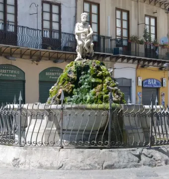 The Fountain of the Genius of Fieravecchia in a city square, with a bearded marble statue on a moss-covered rocky mound behind a protective metal fence.