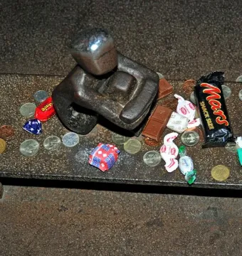 Järnpojke sculpture with a shiny head on a raised platform covered with coins, candies, chocolate bars, and a yellow flower.