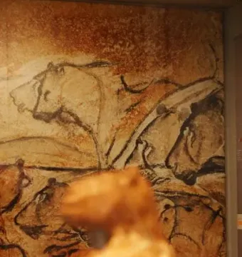 Blurred Lion-man sculpture in the foreground with cave-painting-style animal figures on the wall behind it.