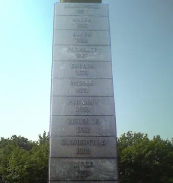 Stone inscription pylon of the Polish Cavalry Monument showing engraved battle names and years against a blue sky.