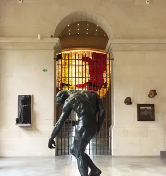 The Three Shades bronze sculpture shown in a museum gallery with a bowed male figure in the foreground.
