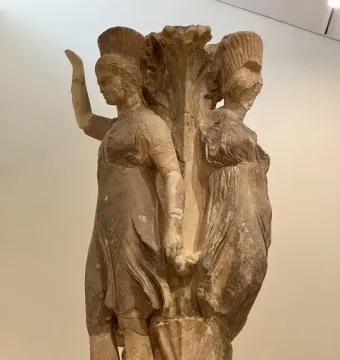 Weathered stone Dancers of Delphi with two draped female figures around a central plant-like column.