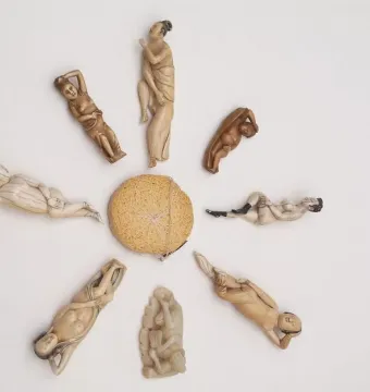 Nine carved diagnostic dolls arranged in a circle around a woven spherical object, with figures in pale ivory and darker tones.