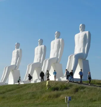 The Man by the Sea sculpture with four white seated figures on a grassy hill and visitors walking below.