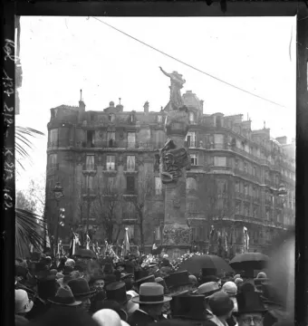 The Monument to Mickiewicz stands in a crowded Paris square during its inauguration, with a statue atop a decorated column and spectators in hats and umbrellas below.