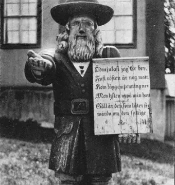 Rosenbom, a wooden statue of a bearded man in a broad-brimmed hat and coat, holding a sign and extending one hand.