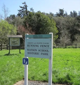 Informational sign for Running Fence in a grassy clearing with a split-rail fence and trees behind it.