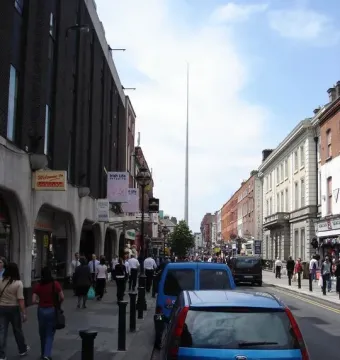 The Spire rises above distant buildings along a busy street with pedestrians and parked cars.