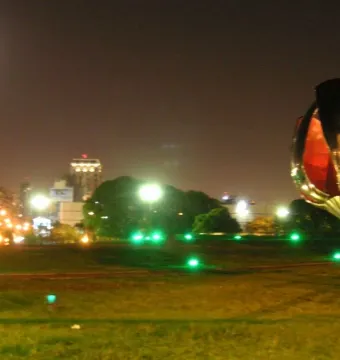 Floralis Genérica partially closed at night, lit by green ground lights with city lights and a bright moon behind it.