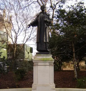 Pankhurst Memorial statue on a tiered stone pedestal in Victoria Tower Gardens, with trees and a low wall behind it.