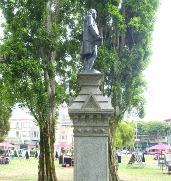 Benjamin Franklin Memorial on a tall stone pedestal in a grassy park with trees and city buildings behind it.