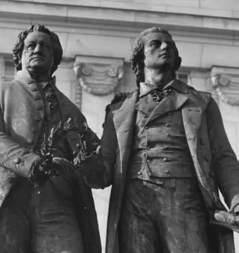 Goethe and Schiller Monument in Weimar, showing two bronze figures in period clothing, one holding a laurel wreath and the other resting an arm on his companion\'s shoulder.