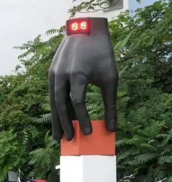 Hand with Clock sculpture with a glowing red digital display, standing on a white pedestal near parked bicycles and a moped.