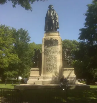 Major General Friedrich Wilhelm von Steuben statue on a tall stone pedestal in a grassy park with trees, with a person sitting at the base behind a low barrier.