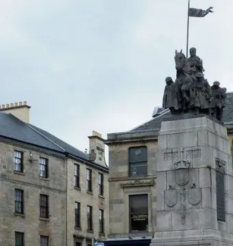 Paisley War Memorial with a stone pillar and dark statue group on horseback, seen above informational boards and a person standing below.