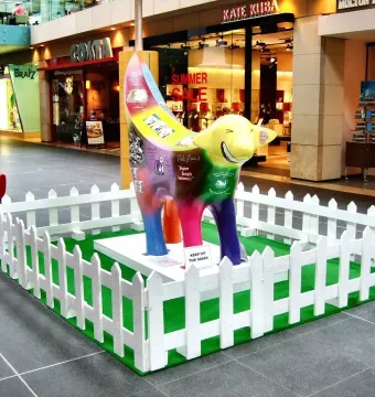 Superlambanana sculpture on artificial grass inside a shopping mall, surrounded by a white picket fence.
