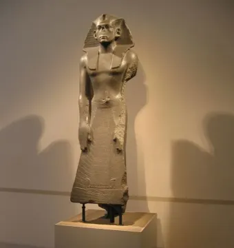 Standing stone statue of Amenemhat III with a striped headdress and long pleated garment.