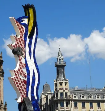 The Face of Barcelona mosaic sculpture rises in front of ornate waterfront buildings under a blue sky.