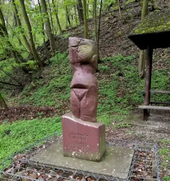 Venus of Petrkovice stone statue on a base beside a gabion wall on a wooded hillside.