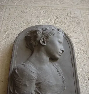 Stone bust of a child in profile, mounted on a light textured wall within an arched frame.
