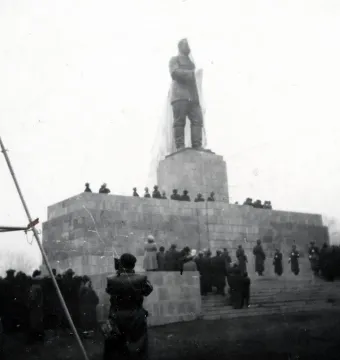 A veiled Stalin monument stands on a tall stone pedestal as a large crowd gathers around it.