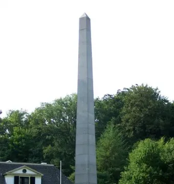 The Needle obelisk stands beside a paved road with a low fence around its base, with trees and a white building in the background.