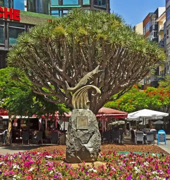 The Chicharro Sculpture, a dark metal fish on a stone pedestal, in front of a large tree and flower bed.