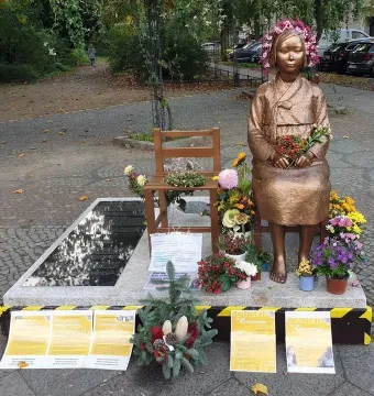 Peace Statue showing a bronze girl seated barefoot on a chair, wearing a flower wreath and holding a small bouquet, surrounded by potted flowers.