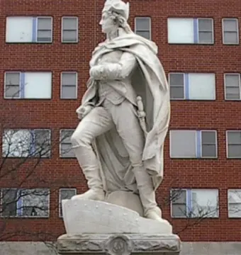 George Washington statue in historical attire standing on a stone pedestal with one foot on a stylized boat.