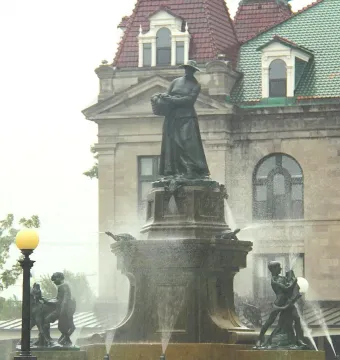 La Fermière bronze statue atop a fountain with water jets, with smaller figures at the base and a historic building behind it.