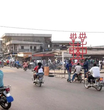 Njé Mo Yé red iron sculpture rising above a busy street with people, motorbikes, and buildings nearby.