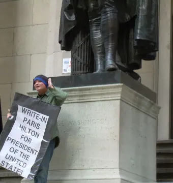 A large George Washington statue stands on a stone pedestal while people gather nearby, including one holding a sign.