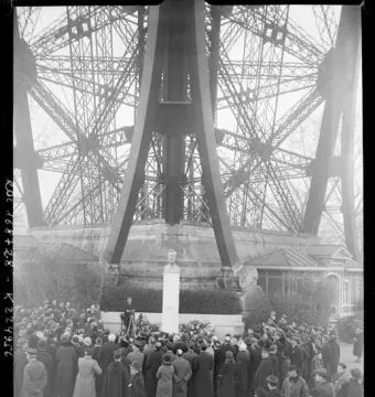 Bust of Gustave Eiffel on a tall pedestal, with floral wreaths and a crowd gathered at the base of the Eiffel Tower.