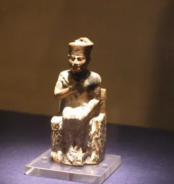 Khufu statuette of a seated figure wearing a tall crown on a clear base.