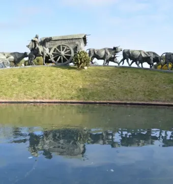 The Monument to the Cart, a bronze sculpture of a covered cart pulled by oxen with a rider on horseback nearby, reflected in a pond.