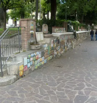 The Muretto of Alassio stone wall decorated with colorful ceramic tiles, a bronze figure perched on top, and a small crest pillar near steps.
