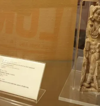 Ivory Lakshmi statuette with elaborate jewelry, headdress, and carved figures at the sides, displayed upright in a clear case.