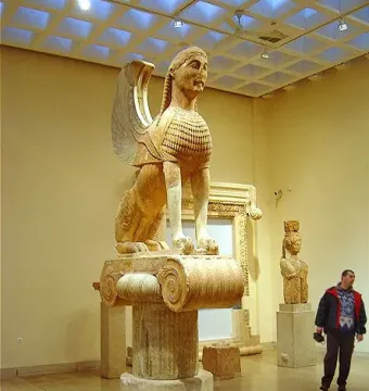 The Sphinx of Naxos statue displayed on a tall column base in a museum hall.